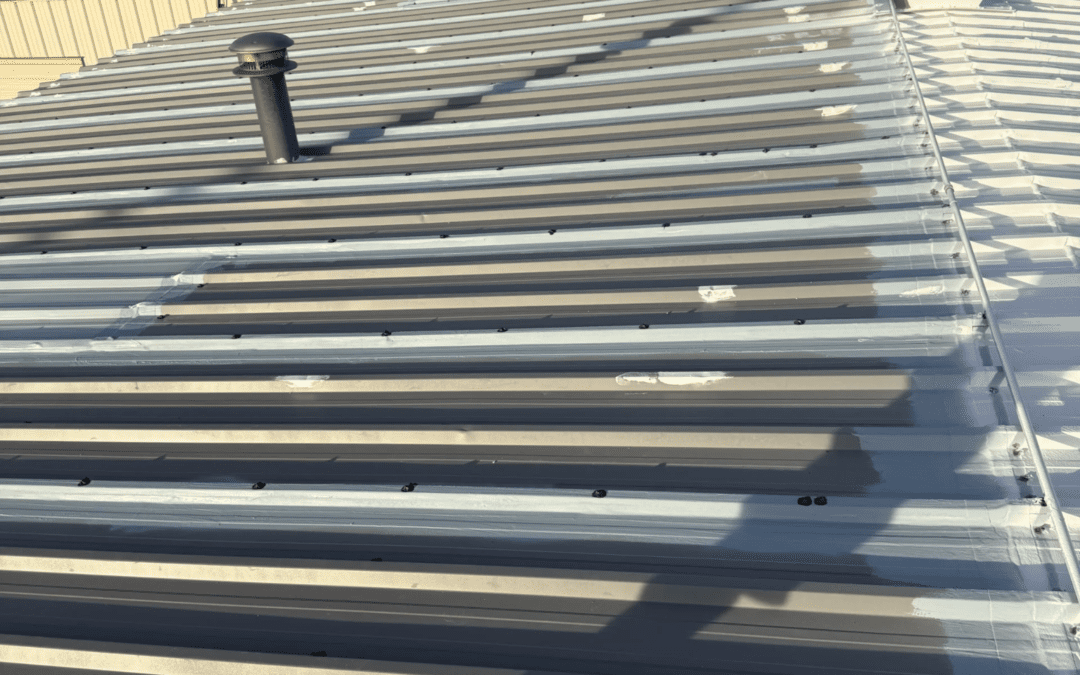 Metal roof with visible seams, bolts, and patches; some pipes and structures protrude from the roof, surrounded by trees and buildings in the background.
