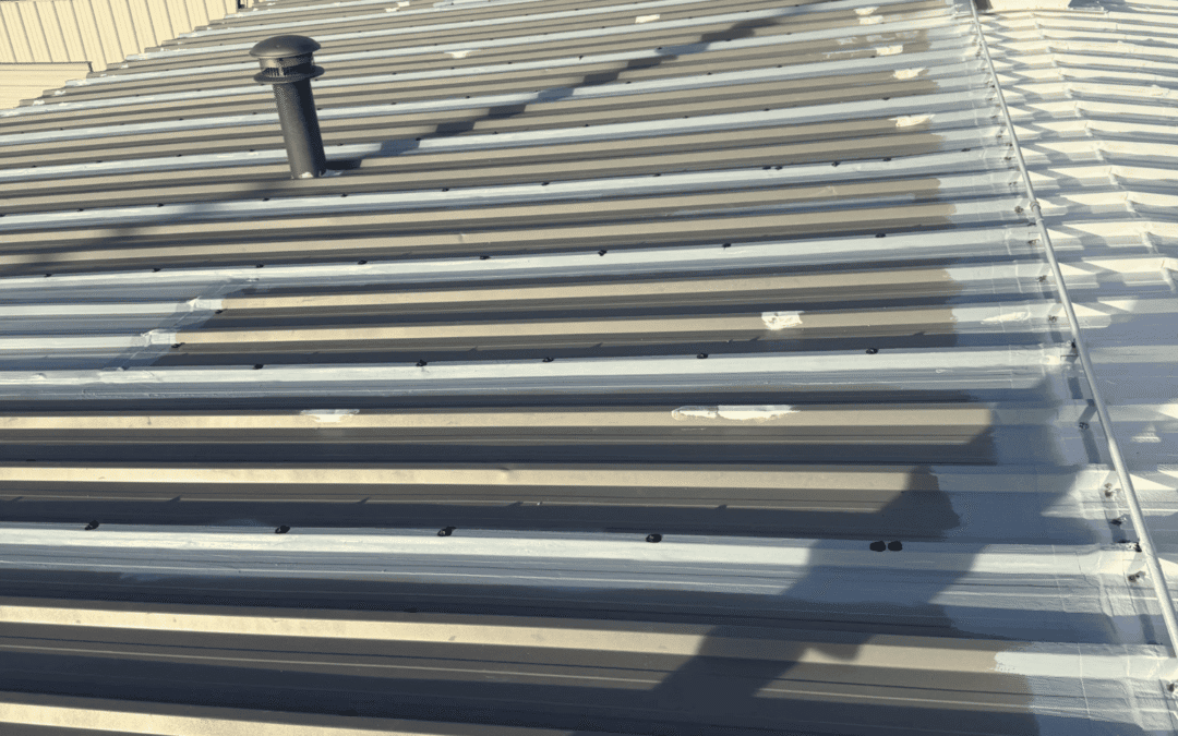 Corrugated metal roof with screws and patches visible, chimney vent, and rooftop shadows; trees and buildings in the background. Reflective roof coatings help lower heat and reduce summer heat load on roofs like this, enhancing comfort and energy efficiency.