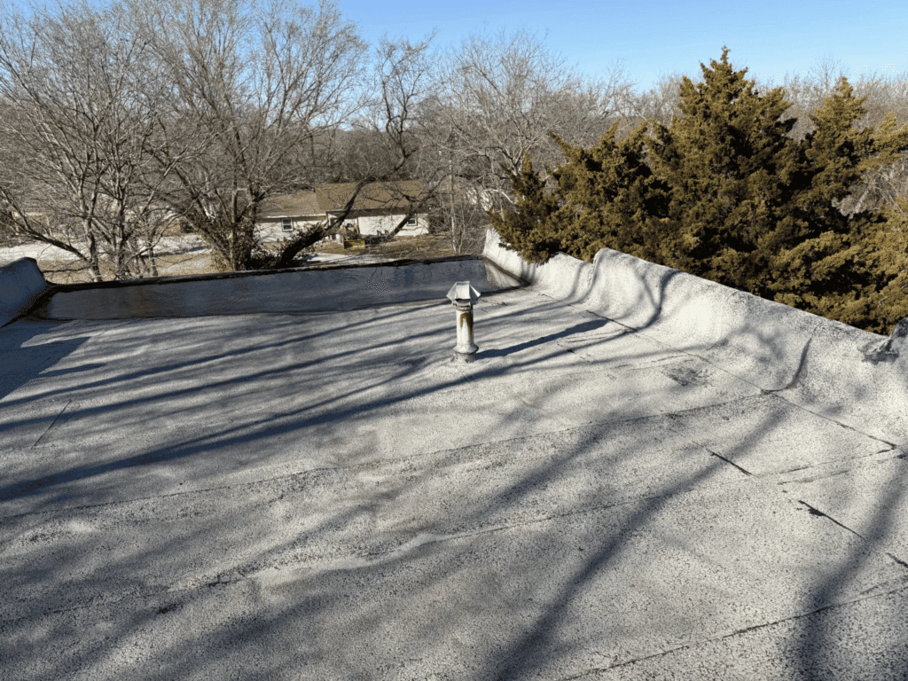 Flat rooftop with a single metal vent in the center, surrounded by leafless trees and some evergreens under a clear blue sky. Reflective roof coatings have been applied to help lower heat and reduce summer heat load, creating a cooler environment beneath the roof.